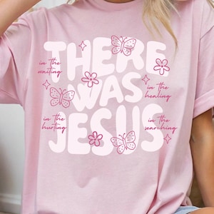 May include: A light pink t-shirt with the words "THERE WAS JESUS" in large, white, bubble letters. The shirt features pink butterflies, flowers, and text that reads "in the waiting, in the hurting, in the healing, in the searching."
