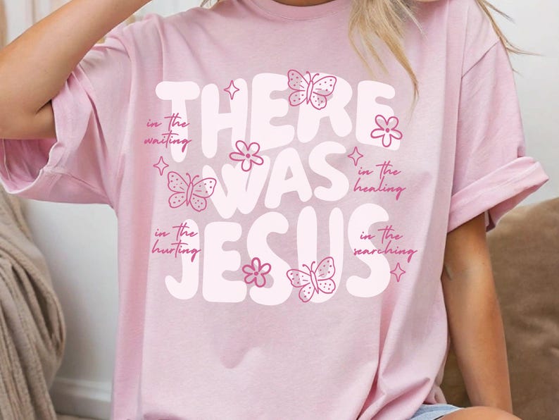 May include: A light pink t-shirt with the words "THERE WAS JESUS" in large, white, bubble letters. The shirt features pink butterflies, flowers, and text that reads "in the waiting, in the hurting, in the healing, in the searching."