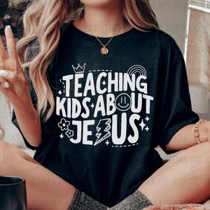 Puede incluir: Camiseta negra con el texto blanco "TEACHING KIDS ABOUT JESUS". El dise&ntilde;o incluye una corona, un arco&iacute;ris, una carita sonriente, un rayo, flores y estrellas. Es una prenda informal.