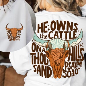 May include: White sweatshirt featuring a brown bull skull graphic with the text "Psalm 50:10" on the front. The back of the sweatshirt displays a larger bull skull design with the words "He owns the cattle on a thousand hills Psalm 50:10".
