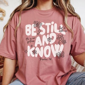 May include: Dusty rose t-shirt with the words "BE STILL AND KNOW" in white, along with floral and butterfly graphics. The shirt also features the phrases "He is our fortress" and "God is with us", and the Bible verse "Psalm 46".