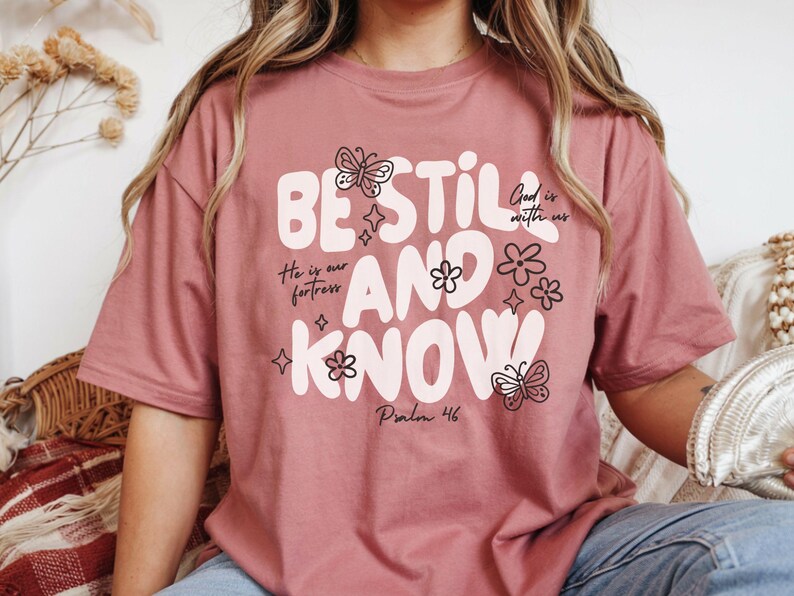 May include: Dusty rose t-shirt with the words "BE STILL AND KNOW" in white, along with floral and butterfly graphics. The shirt also features the phrases "He is our fortress" and "God is with us", and the Bible verse "Psalm 46".