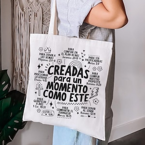 May include: A white canvas tote bag with black text. The text reads "CREADAS para un MOMENTO COMO ESTE" (CREATED for a MOMENT LIKE THIS) surrounded by other phrases and Bible verses. The bag is held by a person.