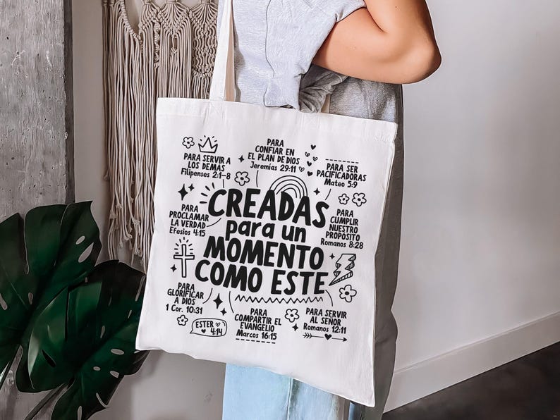 May include: A white canvas tote bag with black text. The text reads "CREADAS para un MOMENTO COMO ESTE" (CREATED for a MOMENT LIKE THIS) surrounded by other phrases and Bible verses. The bag is held by a person.