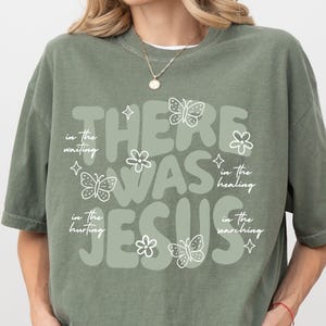 May include: Sage green t-shirt with the text "THERE WAS JESUS" in a rounded font. The design includes white butterflies, flowers, and stars. Additional text reads "in the waiting, in the hurting, in the healing, in the searching."
