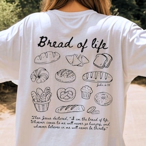May include: White t-shirt with a black illustration of various bread types, including a croissant, pretzel, and loaf. The text "Bread of life" is above the bread images, with a Bible verse below.