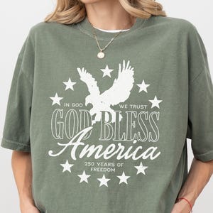 May include: An olive green short-sleeve t-shirt featuring a white graphic of an eagle surrounded by stars. The text reads: IN GOD WE TRUST, GOD BLESS America, and 250 YEARS OF FREEDOM. The design is centered on the chest of the garment.