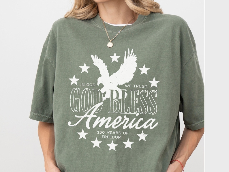 May include: An olive green short-sleeve t-shirt featuring a white graphic of an eagle surrounded by stars. The text reads: IN GOD WE TRUST, GOD BLESS America, and 250 YEARS OF FREEDOM. The design is centered on the chest of the garment.