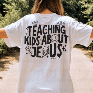 Puede incluir: Camiseta blanca con la frase negra "TEACHING KIDS ABOUT JESUS". El dise&ntilde;o incluye una corona, un arco&iacute;ris, una carita sonriente, flores, un rayo y estrellas. Una camiseta informal.