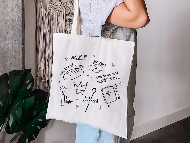May include: White canvas tote bag with black hand-drawn illustrations and text that reads "JESUS IS... the bread of life, the rock, the true vine, the light, Lord, the shepherd, the word".