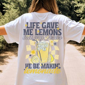 May include: White t-shirt with a graphic design on the back. The design features the text "LIFE GAVE ME LEMONS but my JESUS HE BE MAKIN' lemonade" with a checkered background, a lemonade drink, and lemon slices.