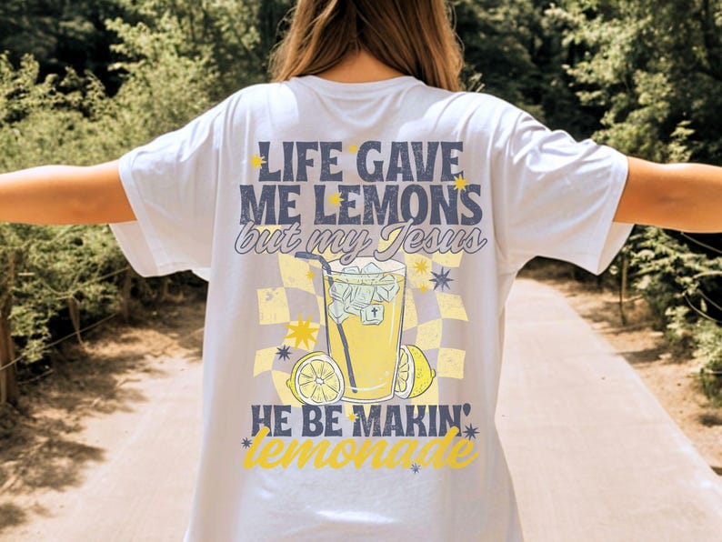 May include: White t-shirt with a graphic design on the back. The design features the text "LIFE GAVE ME LEMONS but my JESUS HE BE MAKIN' lemonade" with a checkered background, a lemonade drink, and lemon slices.