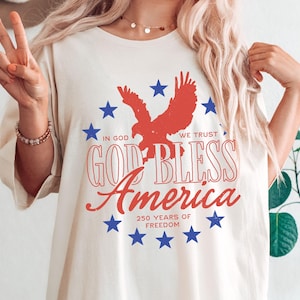 May include: A cream-colored t-shirt featuring a red eagle silhouette surrounded by blue stars. The text reads "IN GOD WE TRUST", "GOD BLESS America", and "250 YEARS OF FREEDOM" in red and white lettering, creating a patriotic graphic design.