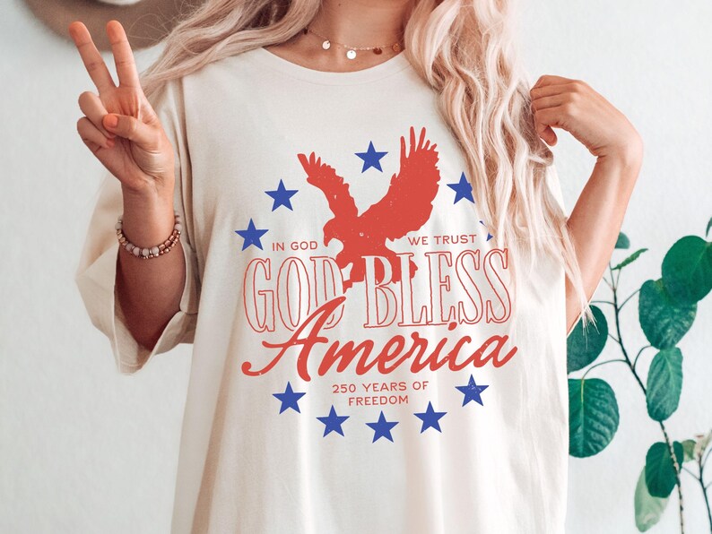 May include: A cream-colored t-shirt featuring a red eagle silhouette surrounded by blue stars. The text reads "IN GOD WE TRUST", "GOD BLESS America", and "250 YEARS OF FREEDOM" in red and white lettering, creating a patriotic graphic design.