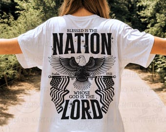 Puede incluir: Camiseta blanca con un diseño gráfico negro que presenta un águila, banderas americanas y el texto "Blessed is the Nation Whose God is the Lord" con una referencia bíblica. El diseño está centrado en la parte posterior de la camiseta.