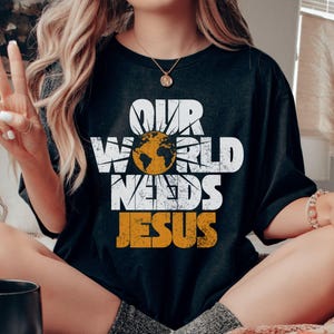 May include: Black t-shirt with the text "OUR WORLD NEEDS JESUS" in white and orange. The design includes a globe inside a sunburst. The shirt is a casual, oversized fit.