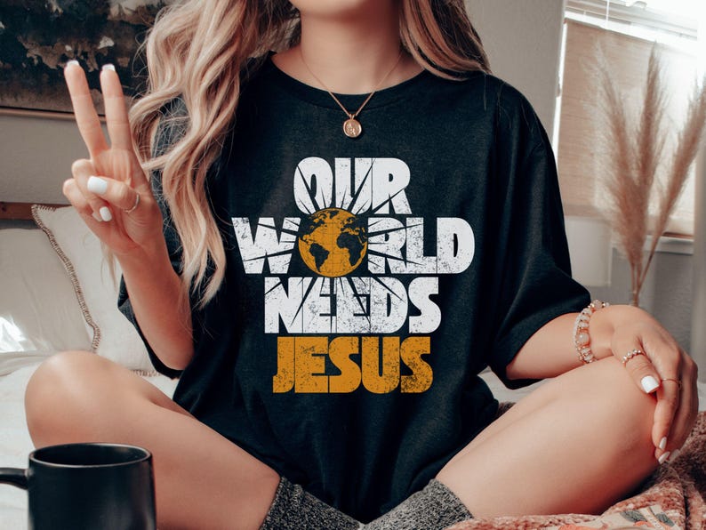 May include: Black t-shirt with the text "OUR WORLD NEEDS JESUS" in white and orange. The design includes a globe inside a sunburst. The shirt is a casual, oversized fit.