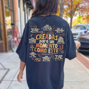 May include: Black t-shirt with colorful text in Spanish, including the phrase "CREADAS para un MOMENTO COMO ESTE." The design features floral and cross motifs, with biblical verses. The shirt is worn outdoors.