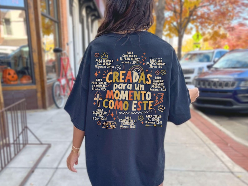 May include: Black t-shirt with colorful text in Spanish, including the phrase "CREADAS para un MOMENTO COMO ESTE." The design features floral and cross motifs, with biblical verses. The shirt is worn outdoors.