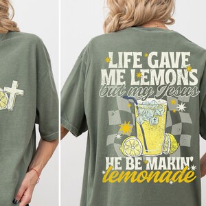 May include: Olive green t-shirt with a cross and lemon graphic on the front. The back features the text "LIFE GAVE ME LEMONS but my Jesus HE BE MAKIN' lemonade" with a lemonade graphic.