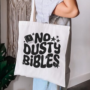 May include: A white tote bag with the phrase "NO DUSTY BIBLES" in large, black, retro-style lettering. The design includes a book icon and stars. The bag is held by a person.