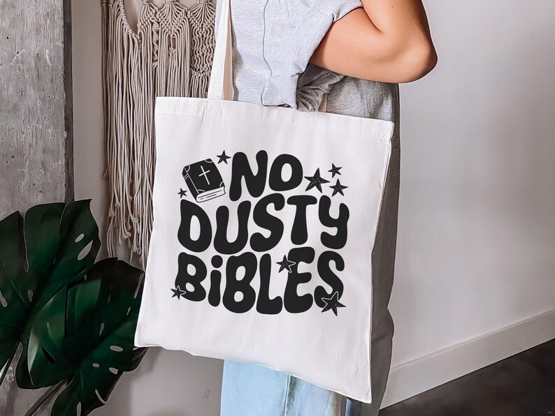 May include: A white tote bag with the phrase "NO DUSTY BIBLES" in large, black, retro-style lettering. The design includes a book icon and stars. The bag is held by a person.