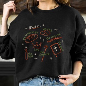 May include: Black sweatshirt with a white graphic design that says "JESUS IS..." and lists several attributes of Jesus, including "the bread of life", "the rock", "the true vine", "the light", "the Lord", "the shepherd", and "the word".