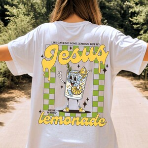 Op de afbeelding: Wit T-shirt met een afbeelding van een cartoon limonadeglas met de tekst "Jesus" en "He be makin' lemonade". Het ontwerp bevat gele en groene accenten en een geruite achtergrond.