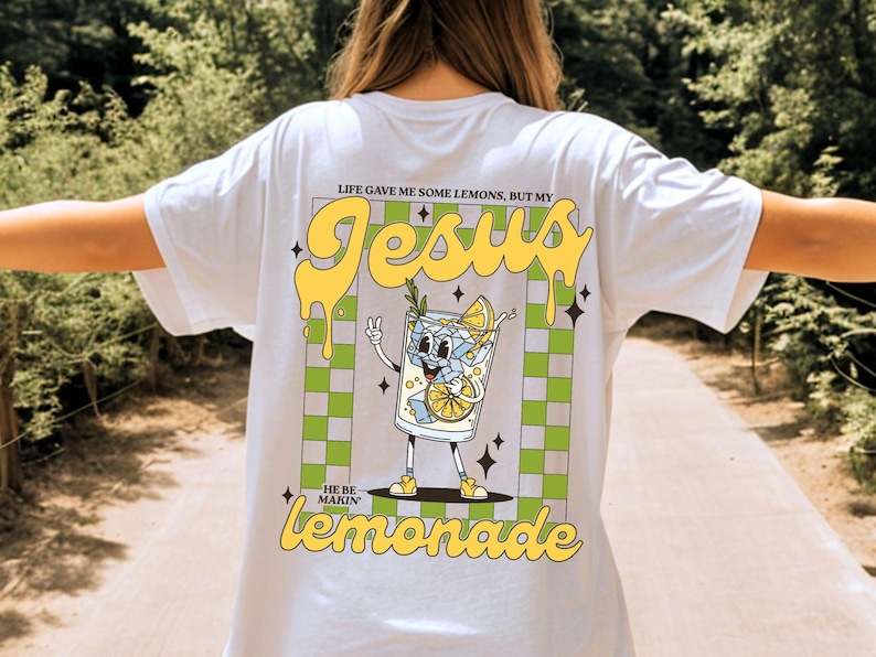 Op de afbeelding: Wit T-shirt met een afbeelding van een cartoon limonadeglas met de tekst "Jesus" en "He be makin' lemonade". Het ontwerp bevat gele en groene accenten en een geruite achtergrond.