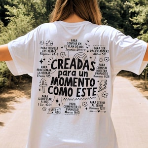 May include: White t-shirt with black text and illustrations. The text reads "CREADAS para un MOMENTO COMO ESTE" (CREATED for a MOMENT LIKE THIS) surrounded by other phrases and Bible verses. The shirt is being worn outdoors.