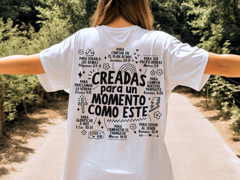 May include: White t-shirt with black text and illustrations. The text reads "CREADAS para un MOMENTO COMO ESTE" (CREATED for a MOMENT LIKE THIS) surrounded by other phrases and Bible verses. The shirt is being worn outdoors.