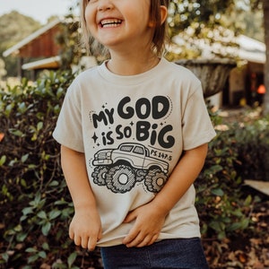 Puede incluir: Un ni&ntilde;o o ni&ntilde;a lleva una camiseta beige con un gr&aacute;fico en blanco y negro de un cami&oacute;n monstruo y el texto "My God is so Big PS.147:5".
