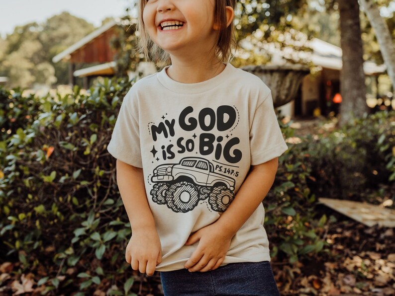 Puede incluir: Un ni&ntilde;o o ni&ntilde;a lleva una camiseta beige con un gr&aacute;fico en blanco y negro de un cami&oacute;n monstruo y el texto "My God is so Big PS.147:5".