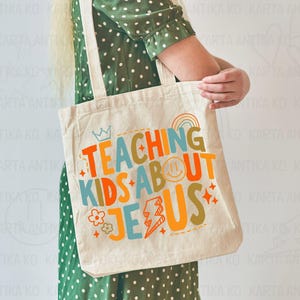 Puede incluir: Bolso tote de color crema con el texto "TEACHING KIDS ABOUT JESUS" en letras coloridas. El dise&ntilde;o incluye una corona, un arco&iacute;ris, una carita sonriente, una flor y un rayo. El bolso tiene dos correas beige.