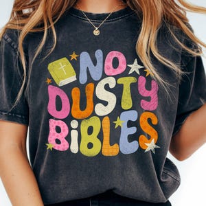 May include: Dark gray t-shirt with the words "NO DUSTY BIBLES" in colorful, retro-style lettering. The design includes a book with a cross and star accents, creating a vintage, inspirational aesthetic.
