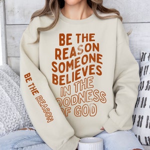 Może przedstawiać: Beżowa bluza z nadrukiem "Be the reason someone believes in the goodness of God" w kolorze pomarańczowym z przodu. Ten sam tekst jest nadrukowany na lewym rękawie w mniejszym rozmiarze.
