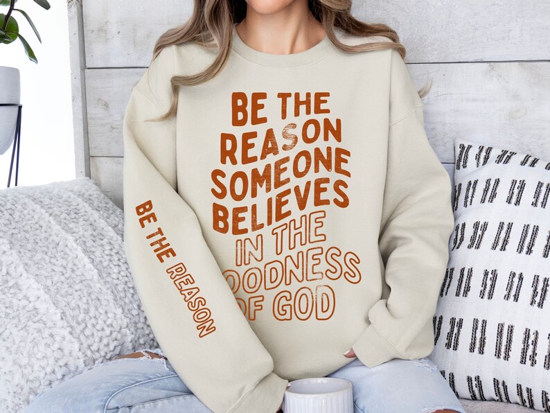 Może przedstawiać: Beżowa bluza z nadrukiem "Be the reason someone believes in the goodness of God" w kolorze pomarańczowym z przodu. Ten sam tekst jest nadrukowany na lewym rękawie w mniejszym rozmiarze.