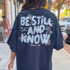 May include: Black t-shirt with the words "BE STILL AND KNOW" in a light blue, wavy font, with floral and butterfly accents. The text "He is our fortress" and "God is with us" are also included. Psalm 46 is written at the bottom.