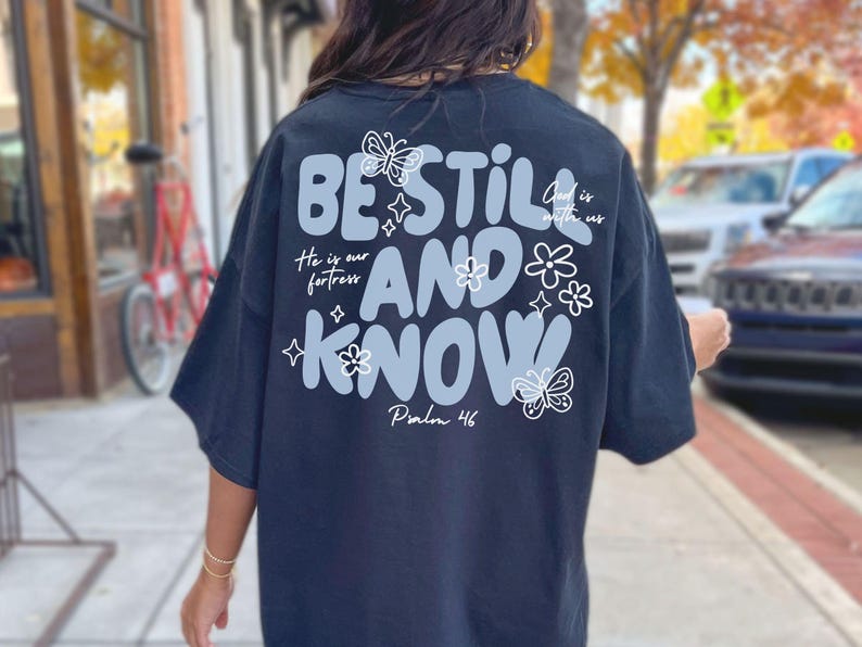 May include: Black t-shirt with the words "BE STILL AND KNOW" in a light blue, wavy font, with floral and butterfly accents. The text "He is our fortress" and "God is with us" are also included. Psalm 46 is written at the bottom.