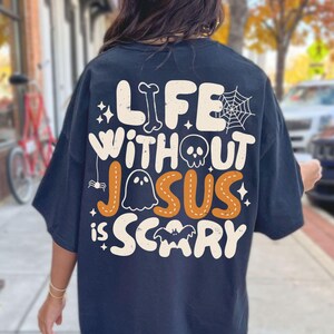 May include: A black t-shirt featuring a large graphic on the back with the text "LIFE WITHOUT JESUS IS SCARY" in cream and orange. The design includes a bone, a spider web, a skull, a ghost, a small spider, and a bat, all in a playful font.