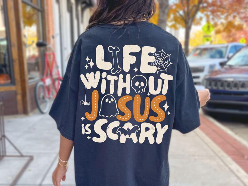 May include: A black t-shirt featuring a large graphic on the back with the text "LIFE WITHOUT JESUS IS SCARY" in cream and orange. The design includes a bone, a spider web, a skull, a ghost, a small spider, and a bat, all in a playful font.