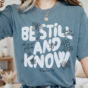 May include: A dusty blue t-shirt with the words "BE STILL AND KNOW" in large white letters. The shirt features butterfly and flower graphics, along with the text "God is with us", "He is our fortress", and "Psalm 46".