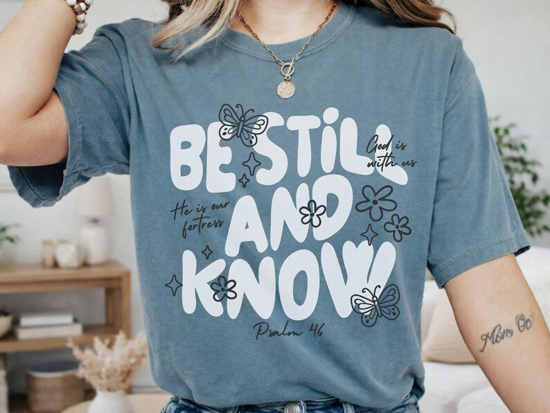 May include: A dusty blue t-shirt with the words "BE STILL AND KNOW" in large white letters. The shirt features butterfly and flower graphics, along with the text "God is with us", "He is our fortress", and "Psalm 46".