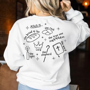 May include: White sweatshirt with black text and illustrations. The text reads "JESUS IS... the bread of life, the rock, the true vine, the Lord, the shepherd, the word". The illustrations include a loaf of bread, a crown, a candle, a rock, a vine, a shepherd's staff, and a book.