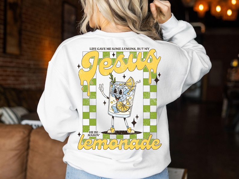 Op de afbeelding: Witte sweatshirt met een afbeelding van een cartoon glas limonade met de tekst "Life gave me some lemons, but my Jesus made lemonade." Het ontwerp bevat een groen-witte geruite achtergrond en gele letters.