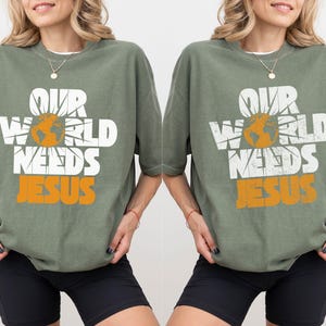 May include: Sage green t-shirt with the text "OUR WORLD NEEDS JESUS" in white and orange. The design includes a globe and sun graphic. The shirt has short sleeves and a relaxed fit.