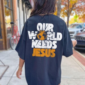 May include: Navy blue t-shirt with the text "OUR WORLD NEEDS JESUS" in white and orange. The design includes a globe and sunburst graphic. The shirt is a casual, oversized fit.
