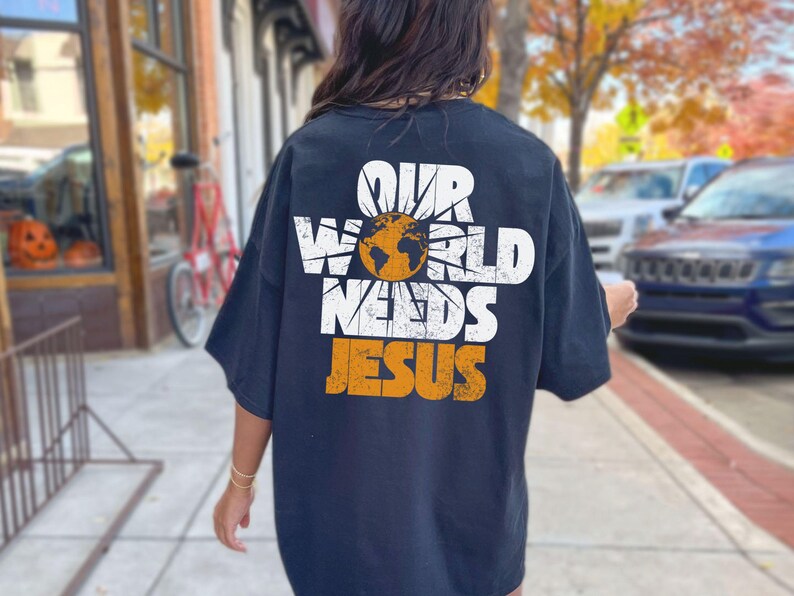 May include: Navy blue t-shirt with the text "OUR WORLD NEEDS JESUS" in white and orange. The design includes a globe and sunburst graphic. The shirt is a casual, oversized fit.