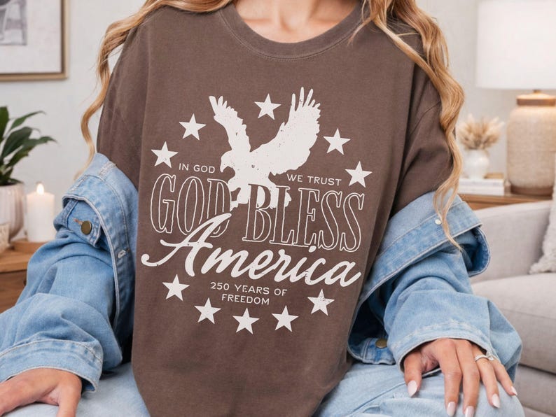 May include: A brown crewneck t-shirt featuring a white graphic of an eagle surrounded by stars. The text reads: IN GOD WE TRUST, GOD BLESS AMERICA, 250 YEARS OF FREEDOM. A light blue denim jacket is layered over the t-shirt, which is worn over blue jeans.