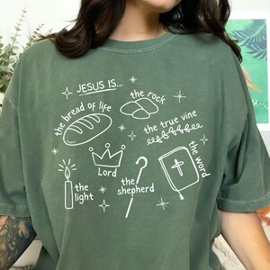 May include: Green t-shirt with white text and illustrations. The text reads "JESUS IS... the bread of life, the rock, the true vine, the word, the light, the shepherd, Lord".
